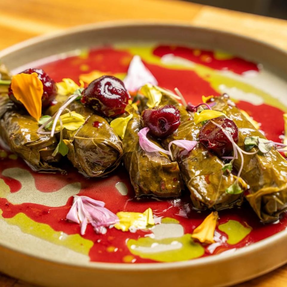 Dolmas, or stuffed grape leaves, garnished with cherries and edible flowers on a plate with red and yellow sauce.
