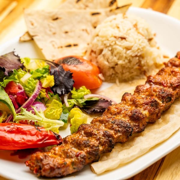 Adana kebab with rice, salad, grilled tomato, and flatbread on a white plate.
