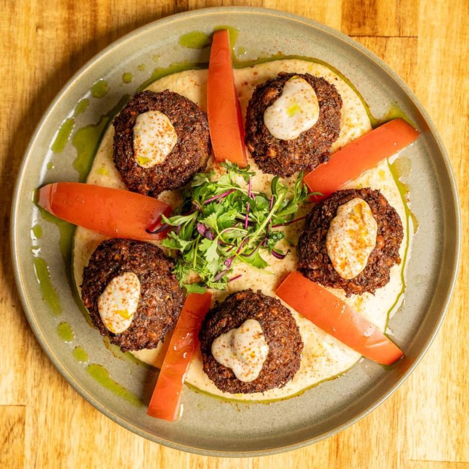 Five falafel patties with white sauce, surrounded by tomato slices and greens on a bed of hummus.