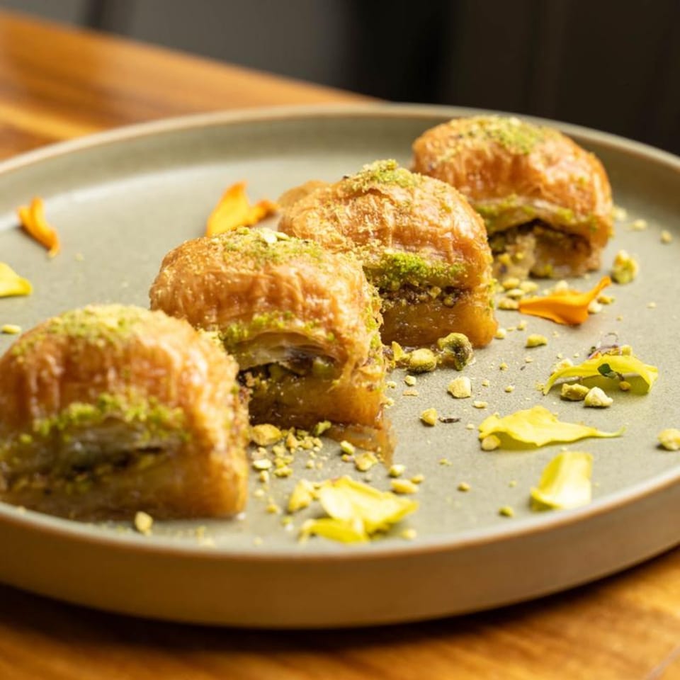 Four pieces of baklava, drizzled with syrup and sprinkled with pistachios, on a grey plate.