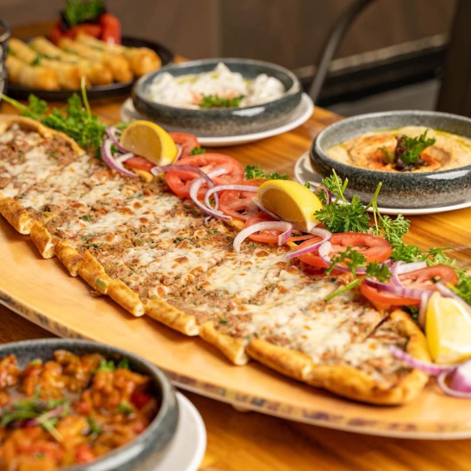 A long, cheesy Mediterranean flatbread with ground meat, tomatoes, onions, and lemons, surrounded by other dishes.