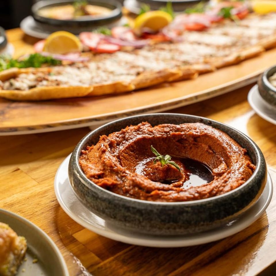 A bowl of red pepper dip with a sprig of herb, and a long flatbread in the background.