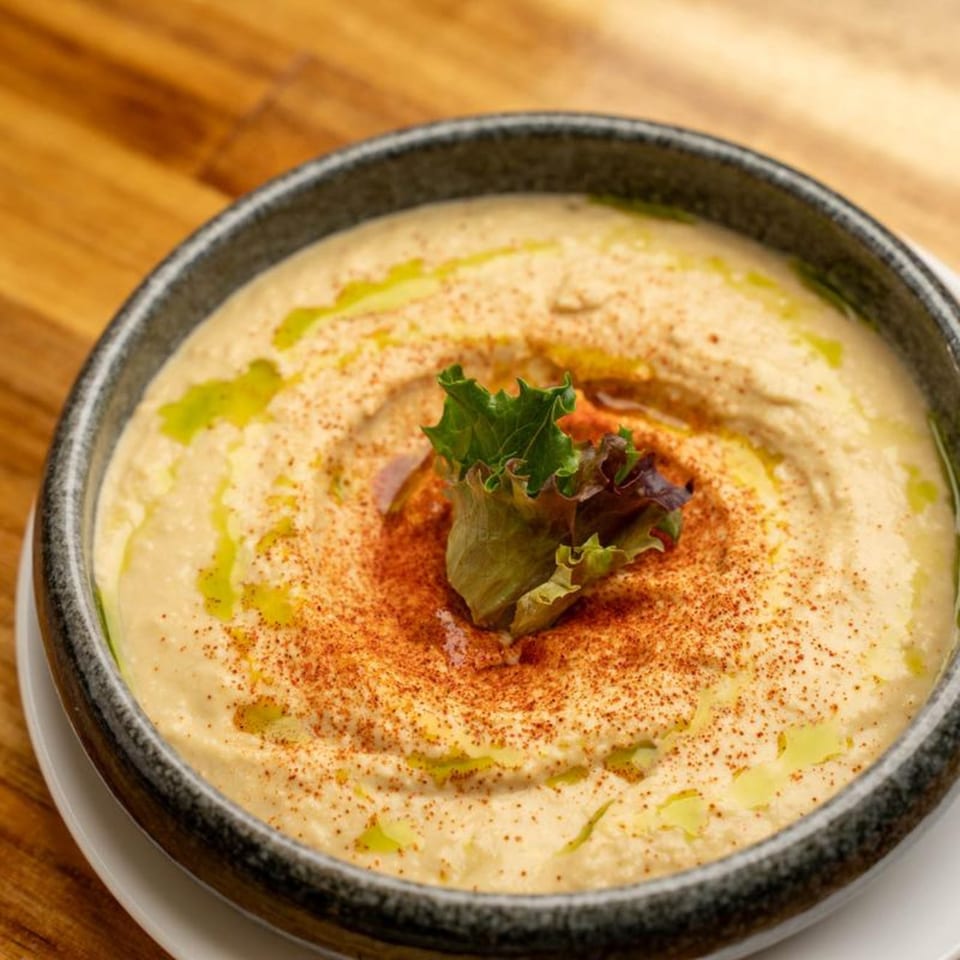 A bowl of creamy hummus, drizzled with olive oil, sprinkled with paprika, and garnished with lettuce.