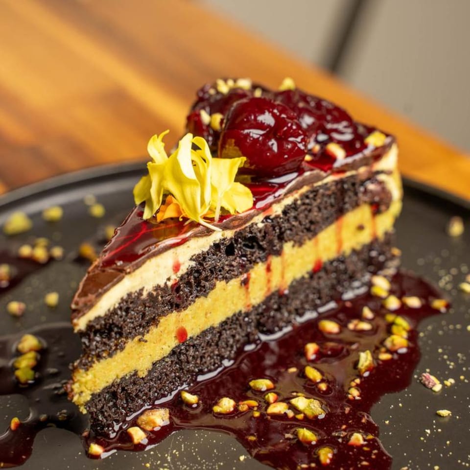 A slice of chocolate cake with yellow filling, cherry topping, and pistachio crumbles on a dark plate.