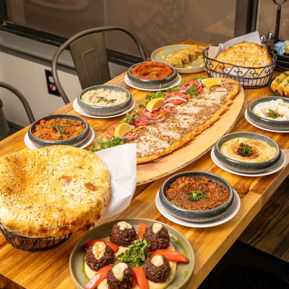 A wooden table laden with various Mediterranean dishes, including flatbread, dips, and a long pide.