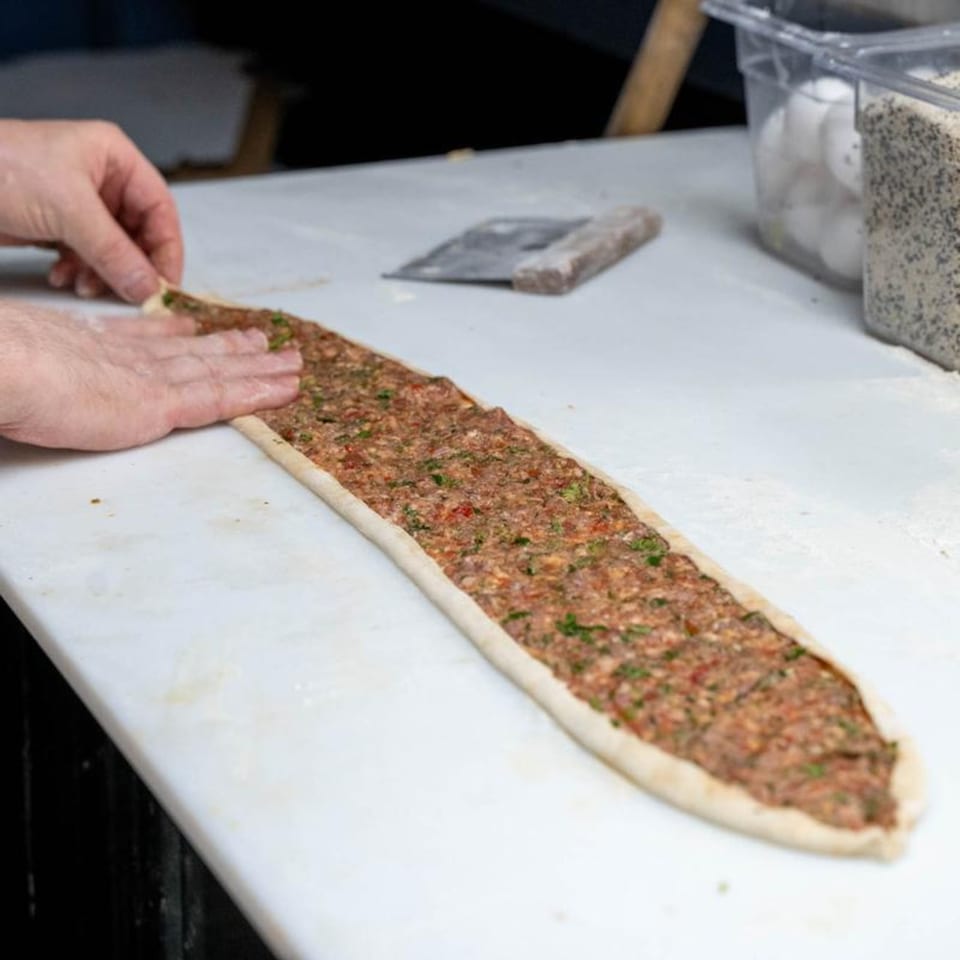 Hands preparing a long, boat-shaped Mediterranean flatbread (pide) filled with ground meat and herbs.