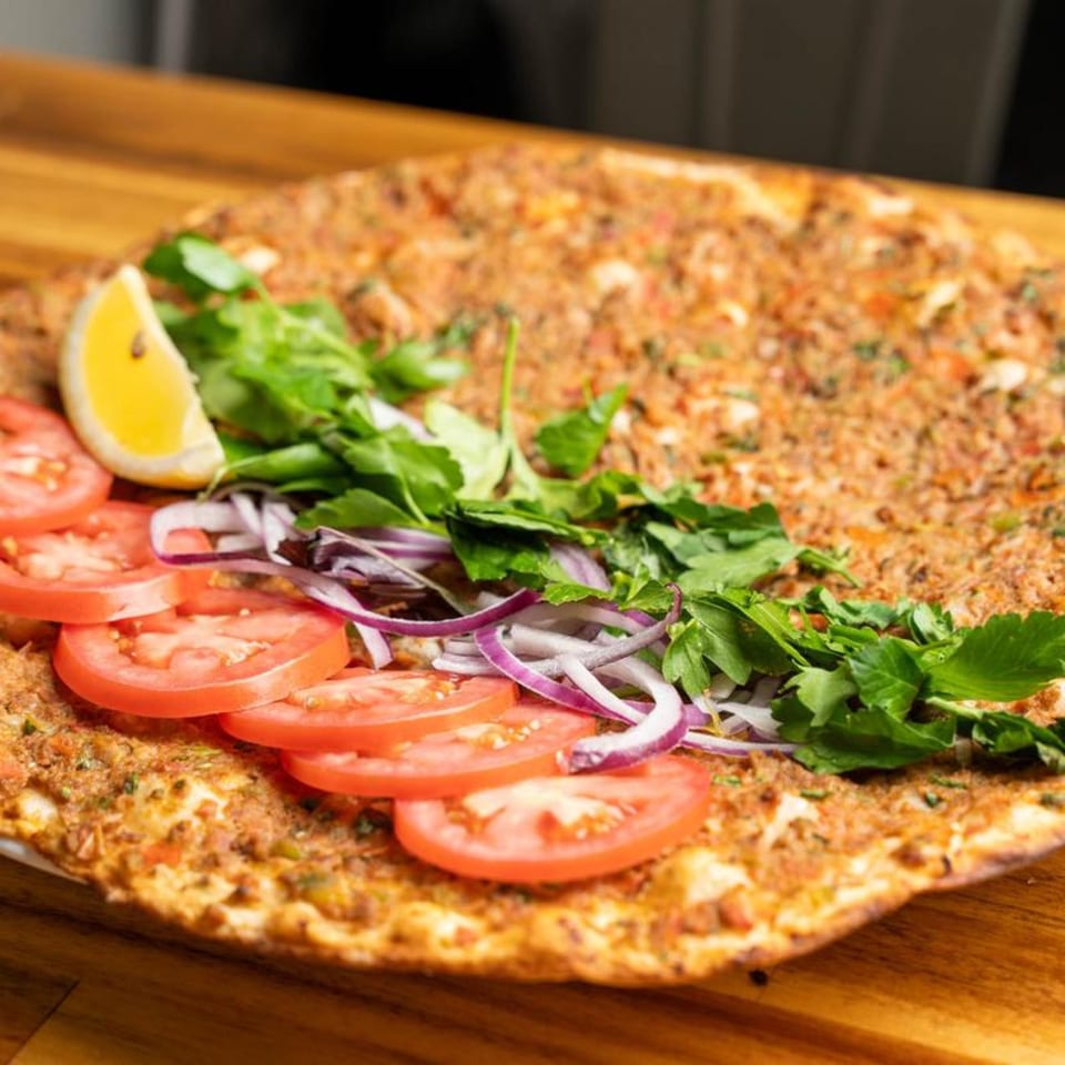 Lahmacun topped with fresh tomatoes, red onions, parsley, and a lemon wedge on a wooden board.