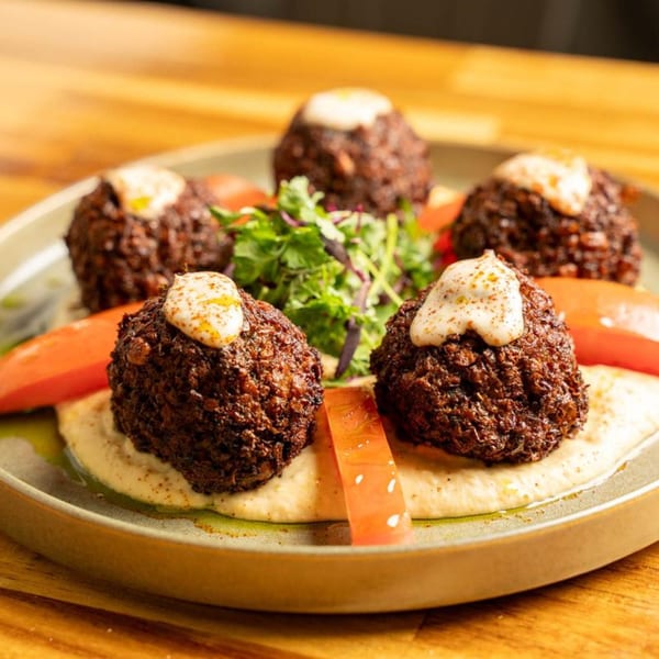Five falafel balls with white sauce, hummus, tomato slices, and greens on a plate.