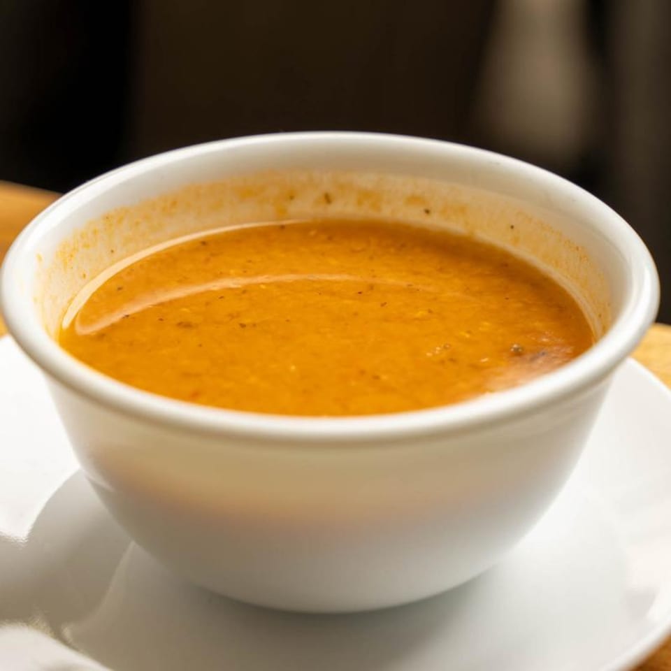A white bowl filled with a rich, orange-colored Mediterranean soup, served on a white saucer.